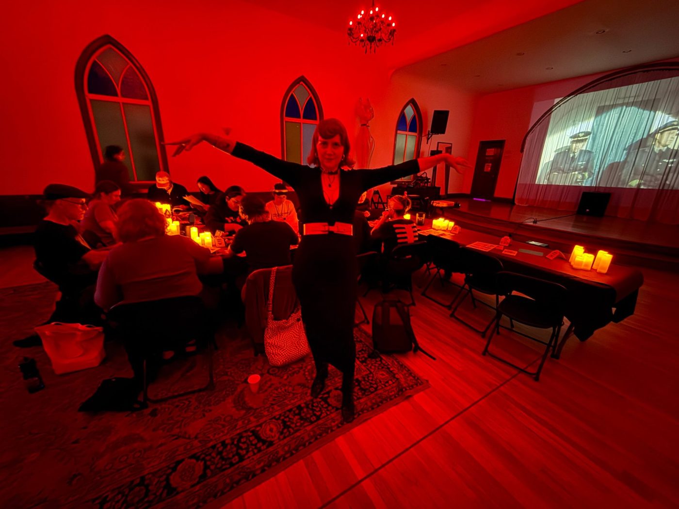 Jem with arms outstretched at Glue Stick Social: a red-lit hall with arched windows, people journaling at candlelit tables, a film on the screen